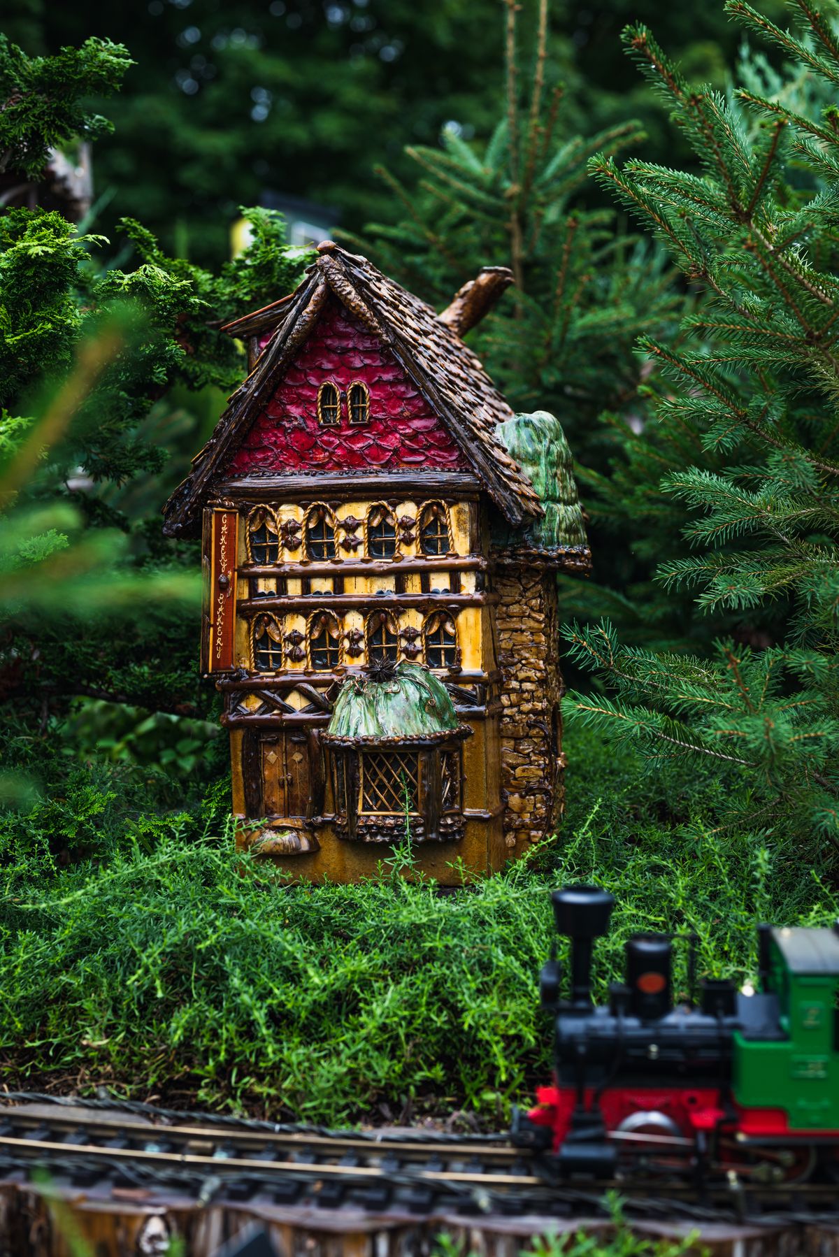Miniature decorative house among green garden plants beside a model railway.